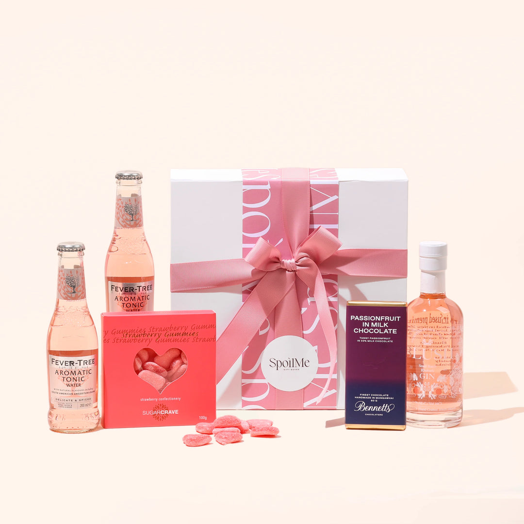 A Blush Gin gift box presentation containing a bottle of blush rhubarb gin, two bottles of tonic, a chocolate bar, and lollies, all tied together with a pink ribbon.