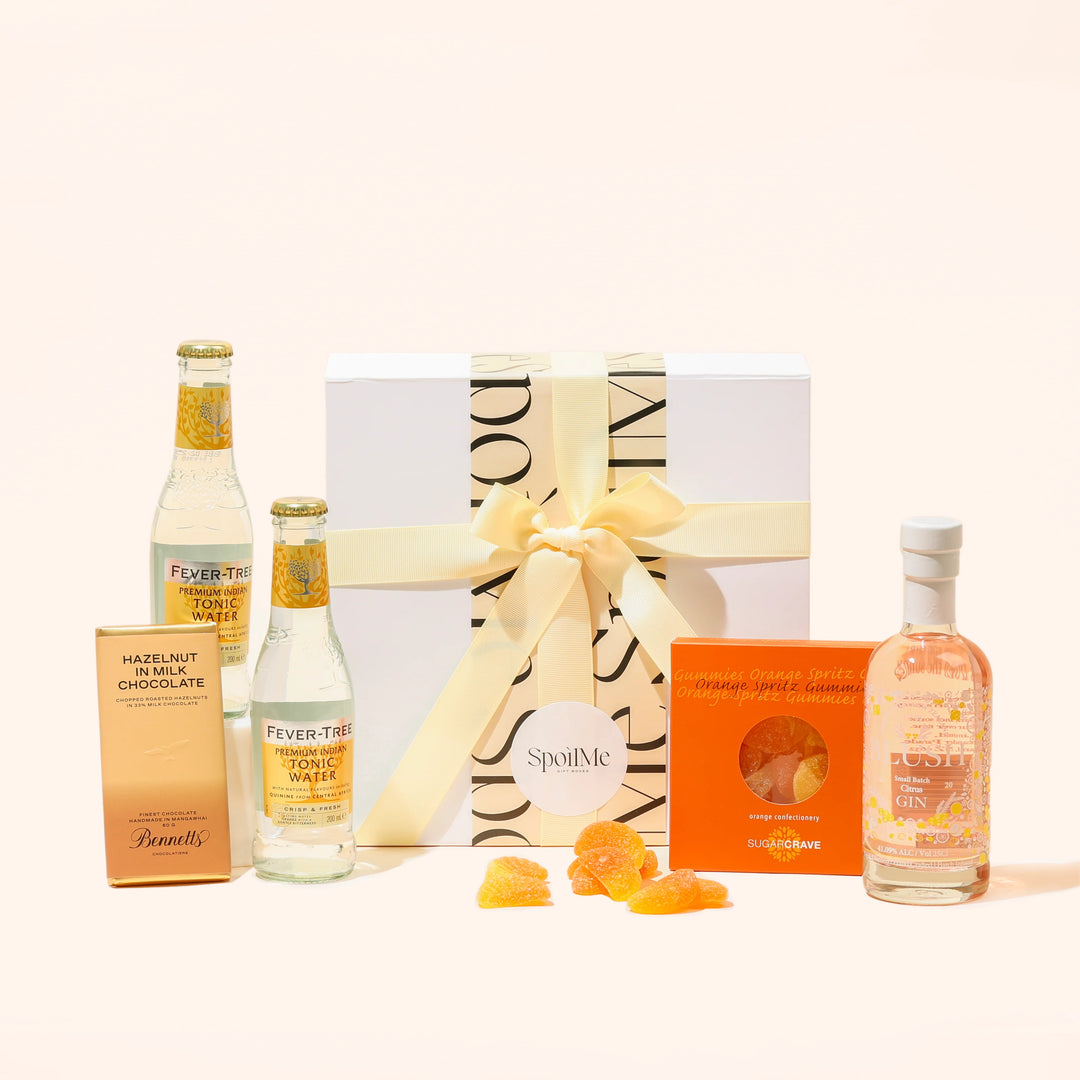 A Blush Gin gift box presentation containing a bottle of Blush citrus pink gin, two bottles of tonic, a chocolate bar, and lollies, all tied together with a pink ribbon.