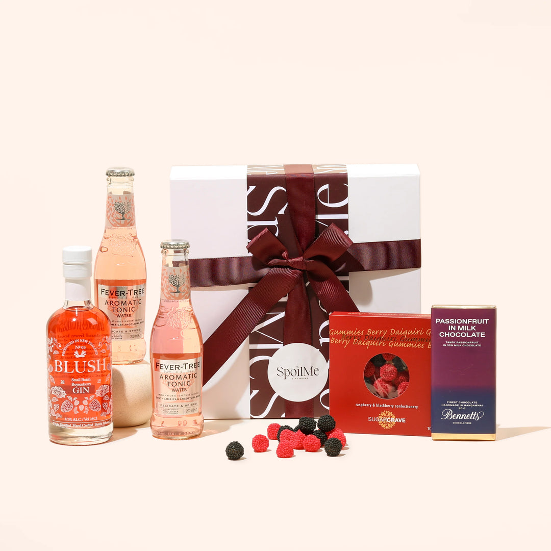A Blush Gin gift box presentation containing a bottle of blush boysenberry gin, two bottles of tonic, a chocolate bar, and lollies, all tied together with a pink ribbon.